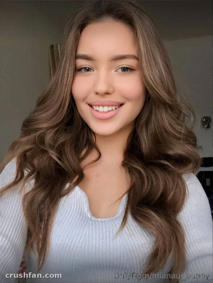 Mia sorrindo com um top canelado branco e longo cabelo ondulado em ambiente interno