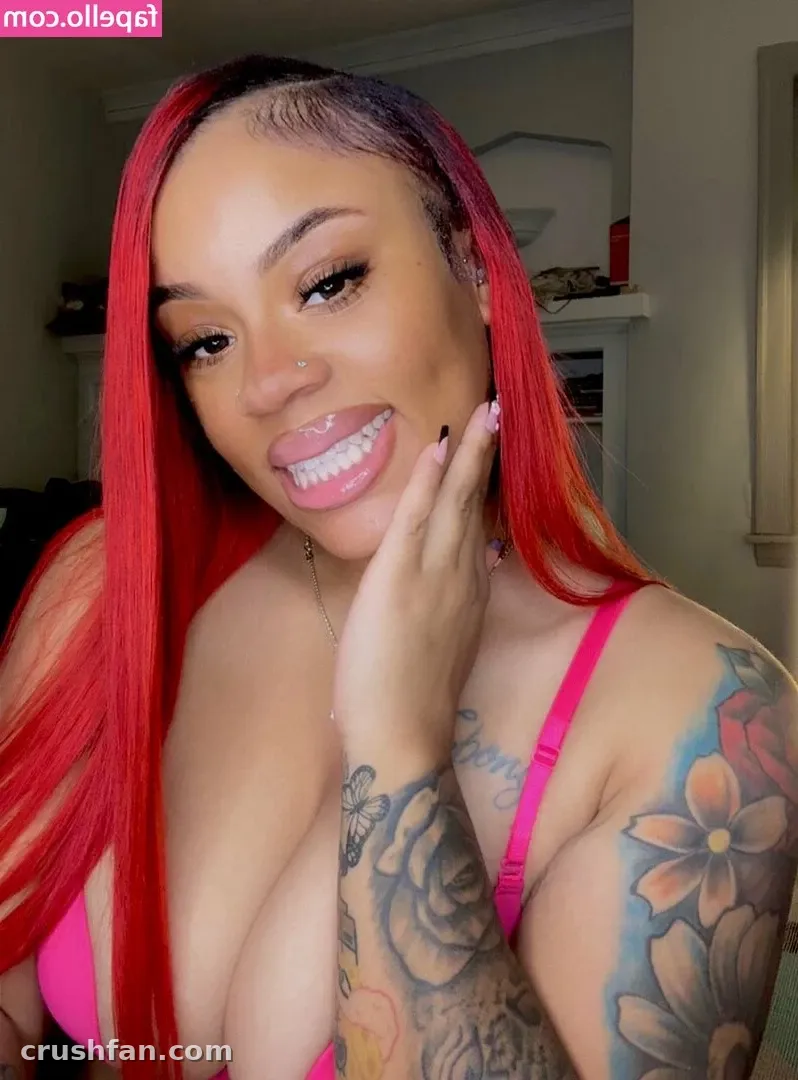 Creator with long red hair and floral arm tattoos smiling in a pink top indoors.