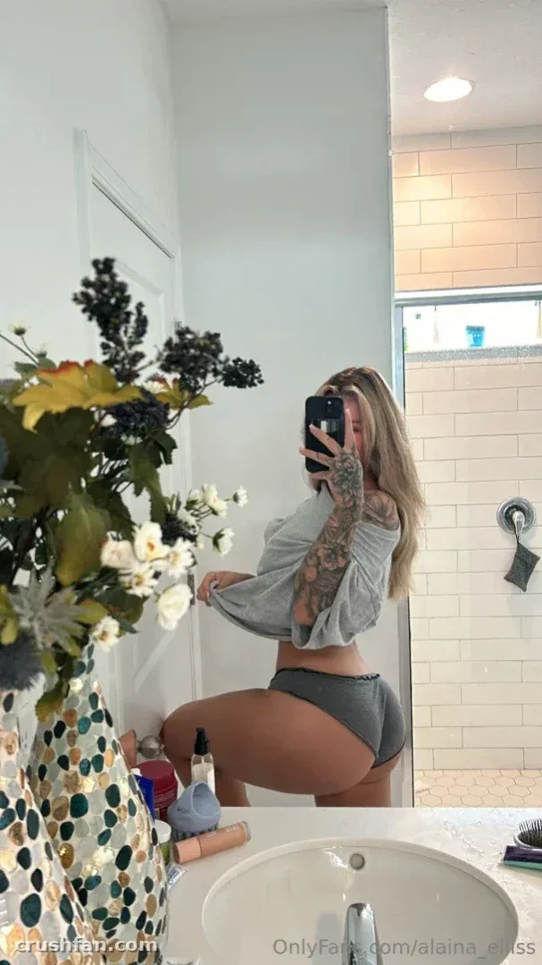 Alaina in a grey top posing for a mirror selfie in a bright bathroom with flowers.