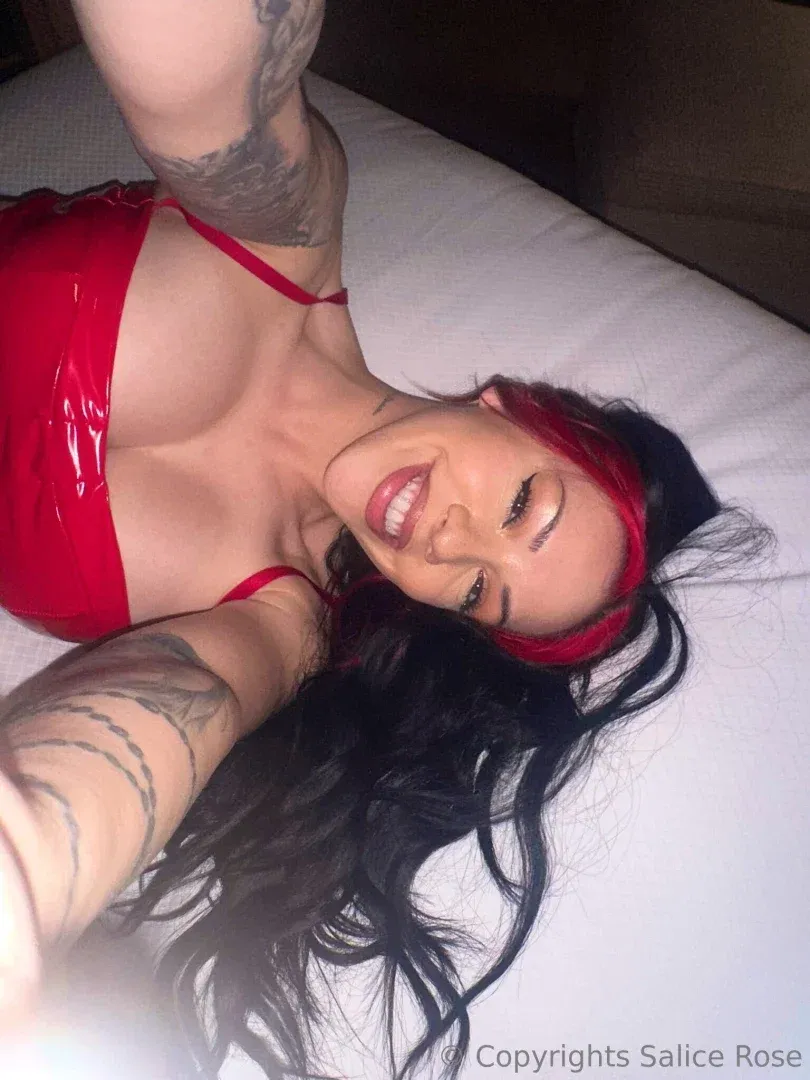 Salice Rose in a glossy red top lying on white bedding with red hair highlights