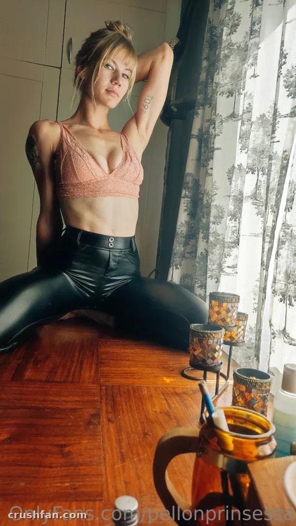 Marjatta seated on a wooden table by a window in a peach bralette and black trousers