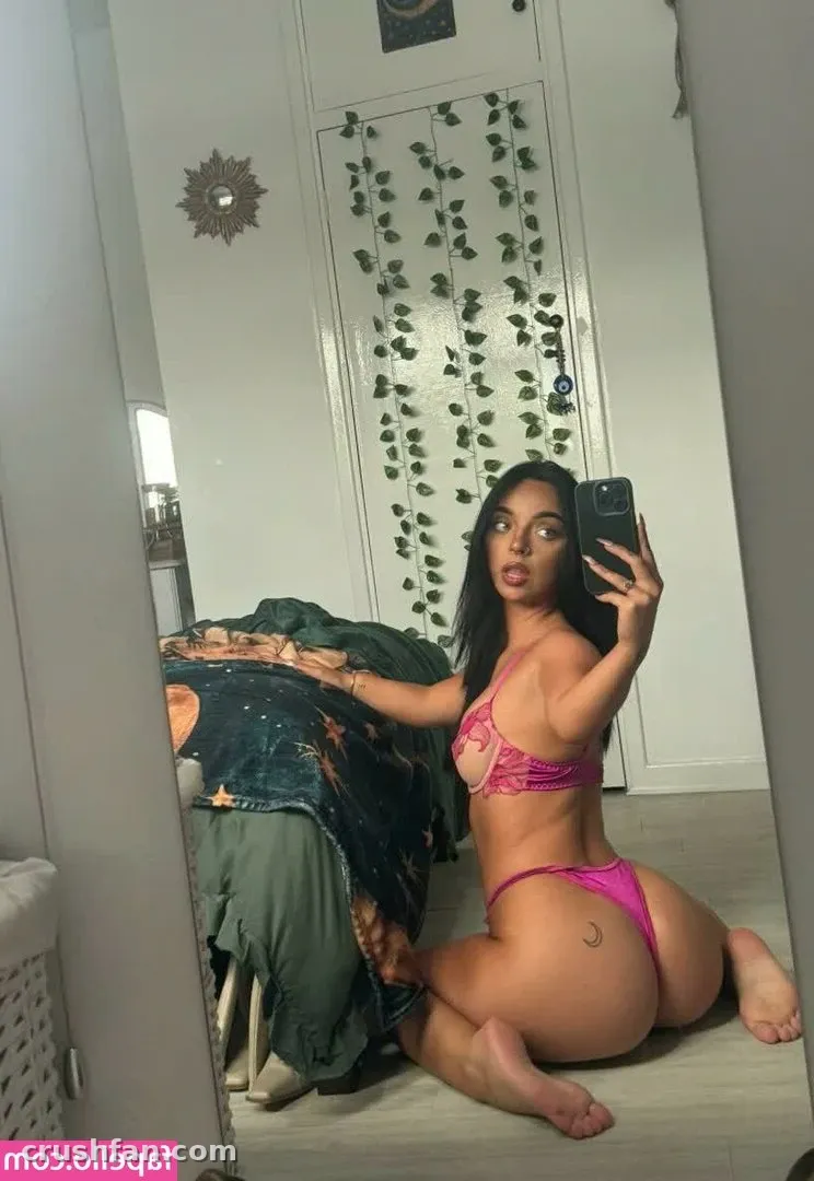 Demi kneeling on a floor taking a mirror selfie in a pink two-piece set, with a bed and ivy-decorated door behind her.