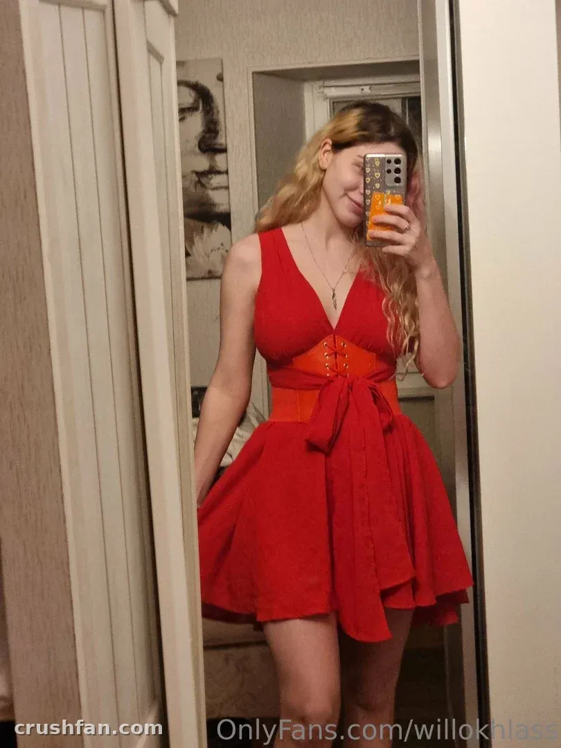 Willokhlass com um vestido vermelho e uma faixa laranja tirando uma selfie no espelho em ambiente interno