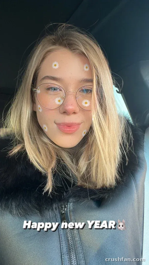 HeyHorny in einem grauen Mantel mit Pelzkragen und Brille, die ein Auto-Selfie mit einem Gänseblümchen-Filter macht.