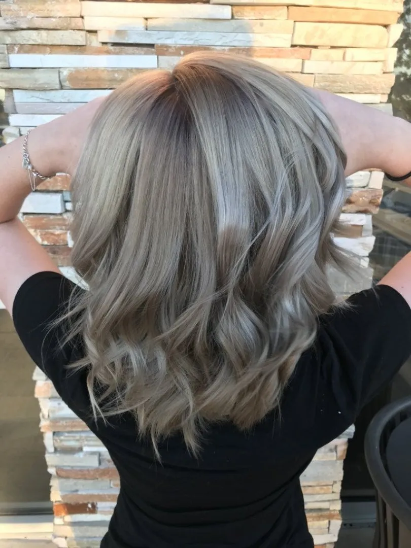Creadora con cabello ondulado rubio ceniza y una blusa negra, vista desde atrás contra una pared de piedra