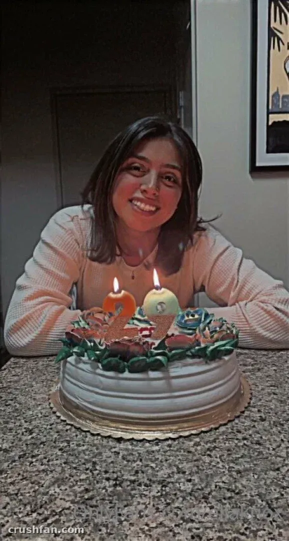 Ella Knox sorrindo atrás de um bolo decorado com velas acesas em um balcão.