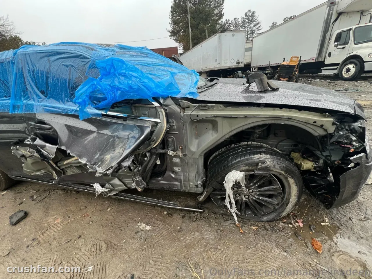 Damaged dark car covered partly with a blue tarp beside white trucks outdoors