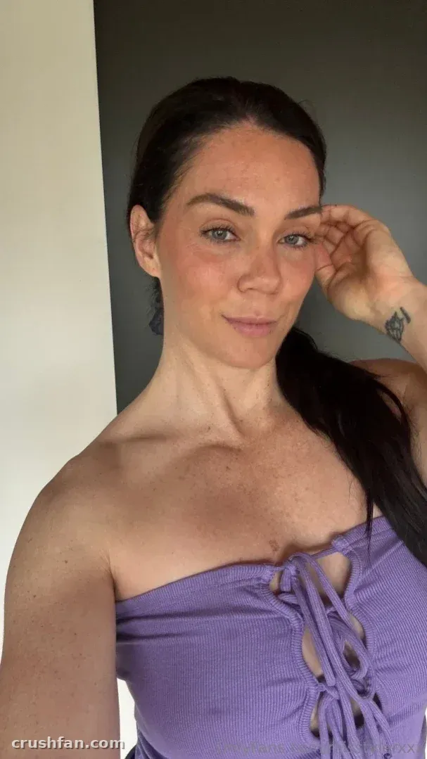 Alison Tyler в сиреневом топе со шнуровкой, рука возле виска, в помещении на фоне однотонной стены.