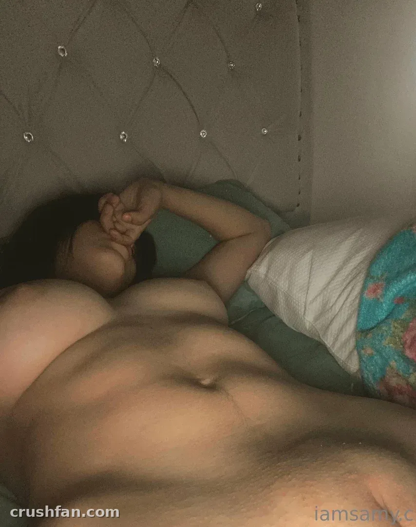 Samy recostada en una cama con cabecero acolchado y ropa de cama verde azulada en luz suave