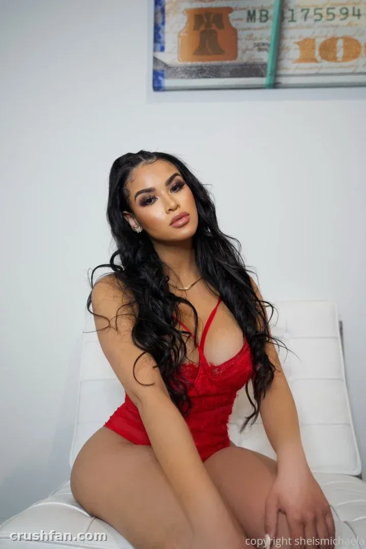 Michaela con un body de encaje rojo sentada en una silla blanca contra una pared lisa