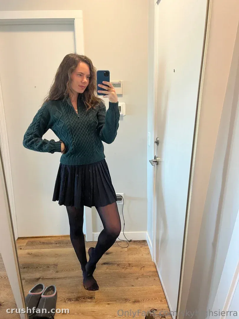 Sierra in einem grünen Pullover und einem schwarzen Faltenrock, die drinnen ein Spiegel-Selfie macht.