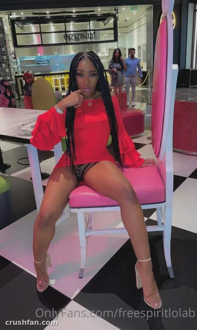 Lola B sitting on a pink chair in a checkered-floor room, wearing a red top and heels.