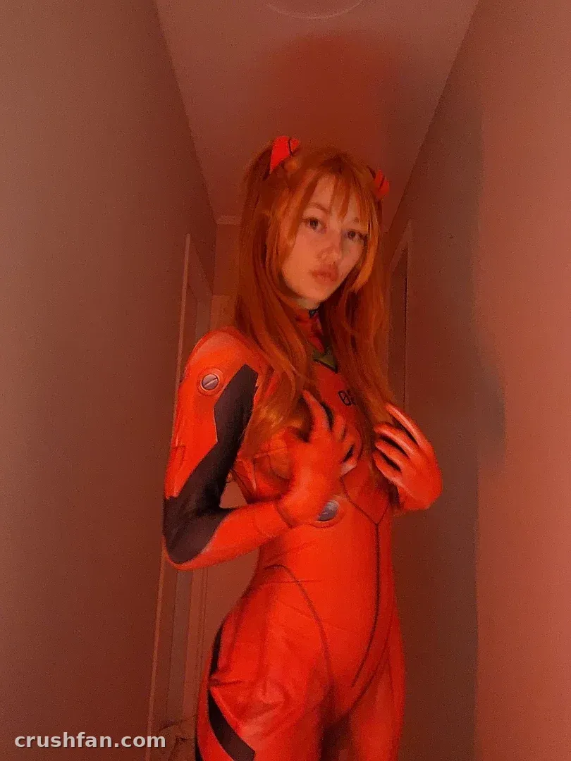Alice NZ em um bodysuit de cosplay vermelho em pé em um corredor com luz suave.
