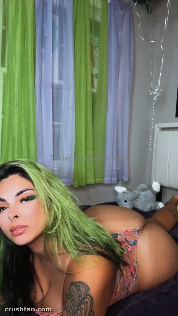 Creator con largo cabello verde recostada en una cama con lencería floral, cortinas verdes y lilas detrás de ella