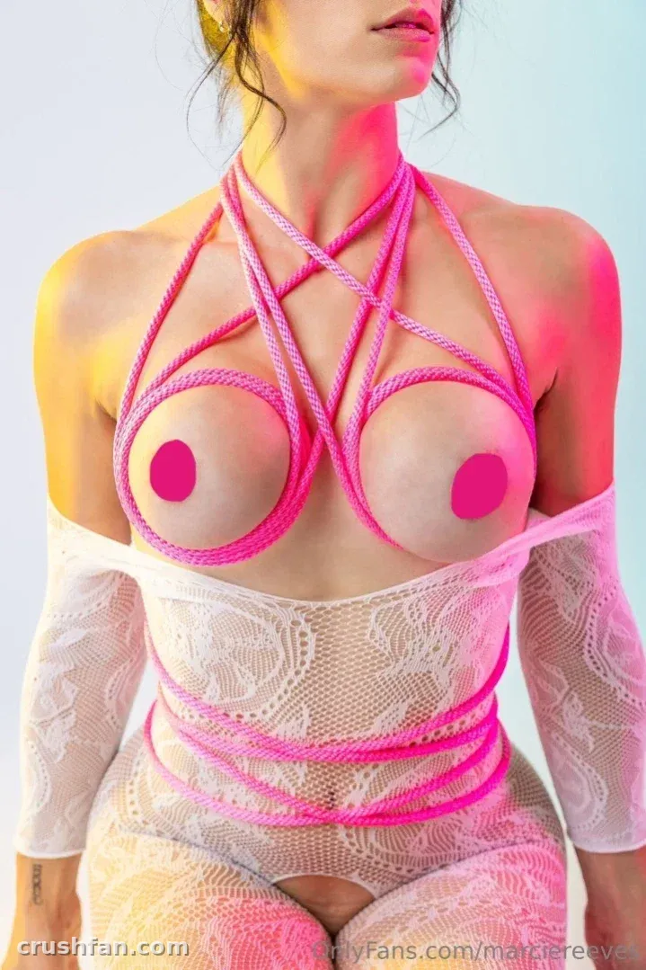 Marcie Reeves portant un body en dentelle blanc et un harnais de corde rose sur un fond uni