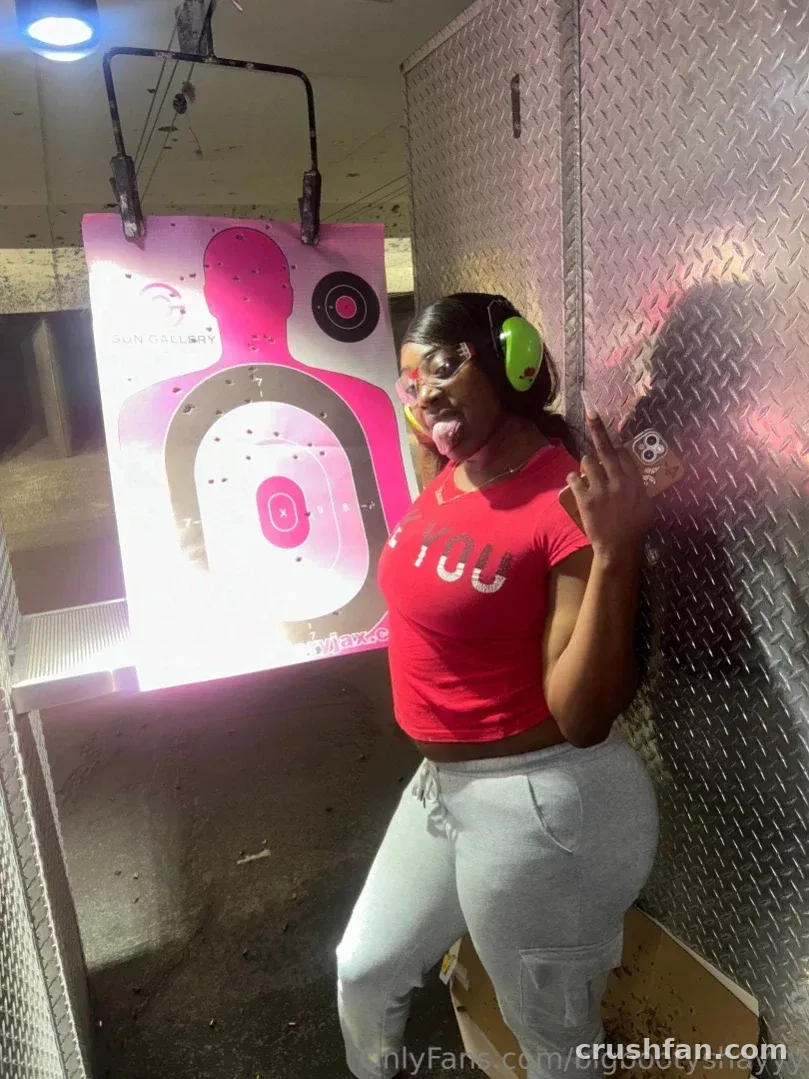 Criadora de top vermelho e calça de moletom cinza ao lado de um alvo rosa em um estande de tiro indoor.