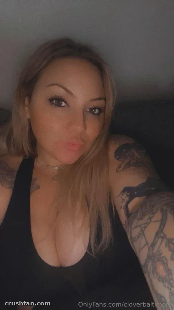 Cloverbaltimore taking a low light selfie in a black top, showing arm tattoos.