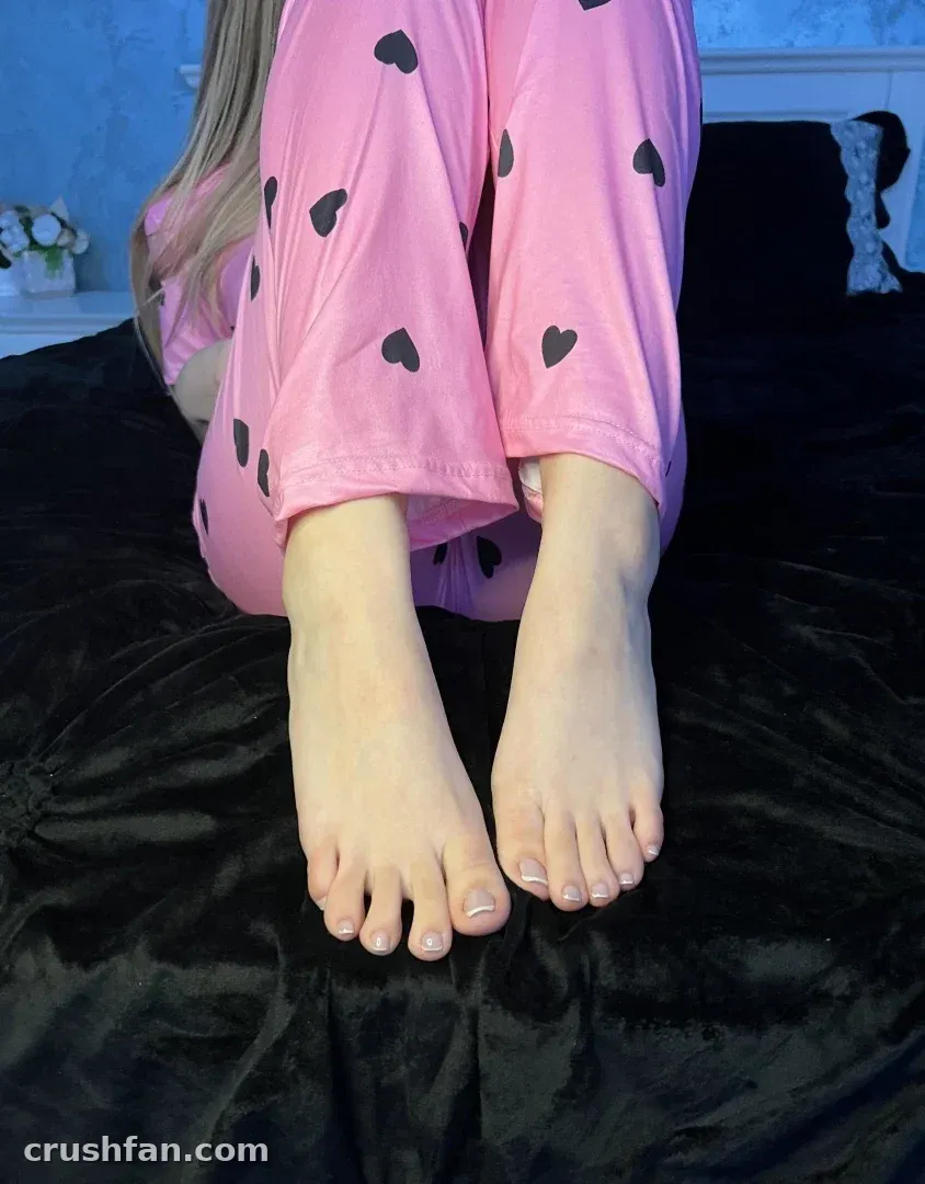 Alexa con ropa de estar por casa rosa estampada de corazones sentada sobre una colcha negra con los pies hacia la cámara