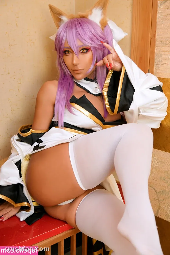 Criadora com peruca roxa e fantasia cosplay branca sentada em um banco vermelho em ambiente interno