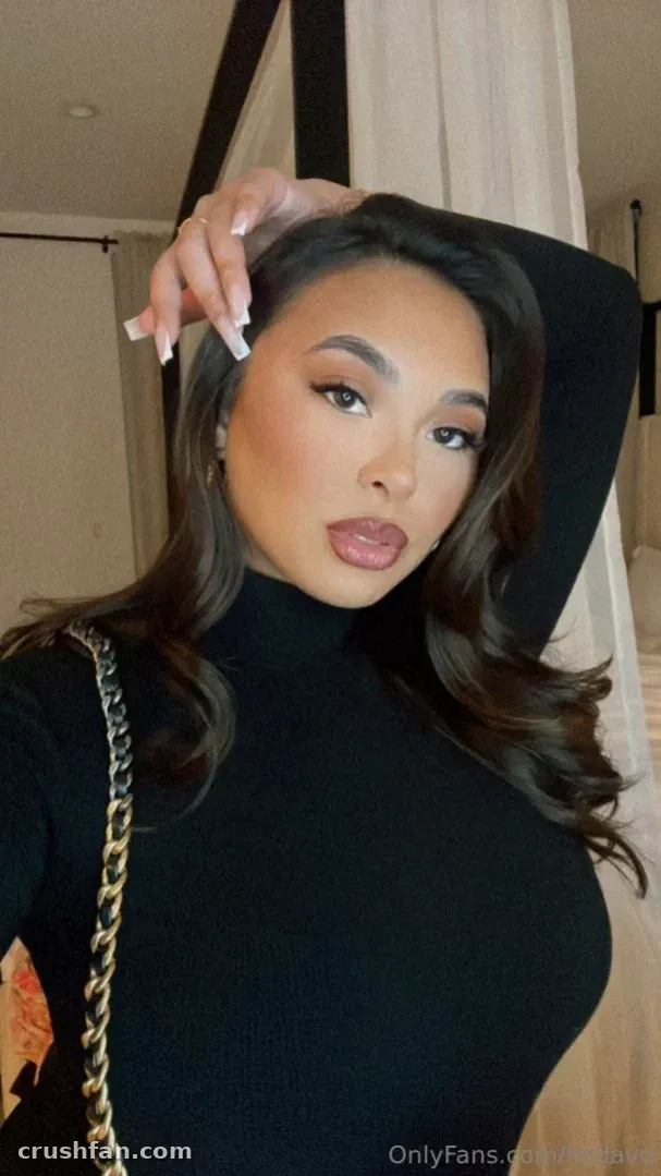 Linda Vo com gola alta preta, cabelo longo e ondulado e alça de corrente dourada