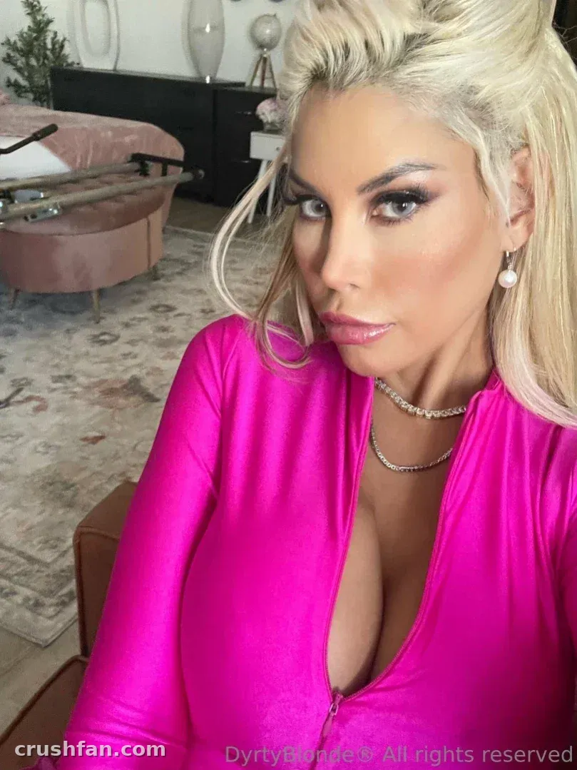 Bridgette B in einem leuchtend pinken Zip-Up-Top drinnen, mit Gliederkette und Perlen-Ohrringen