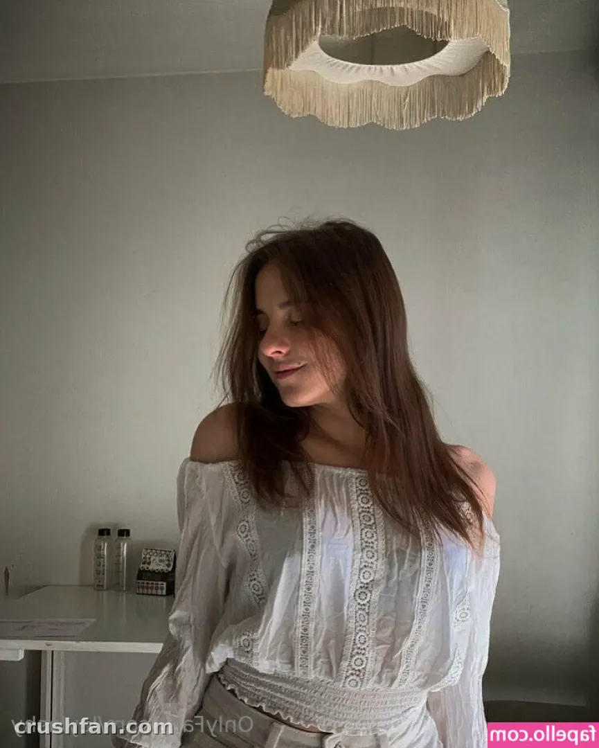 Mulher com uma blusa branca bordada ombro a ombro em pé sob uma luminária pendente de franjas em uma sala iluminada