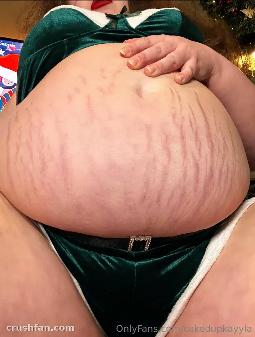 CakedupKayyla in a green velvet holiday outfit with white trim, hand resting on her midsection indoors.