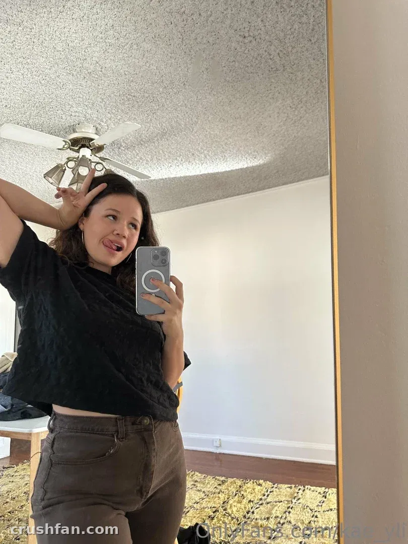 Aka Yhivi in a black top and brown trousers posing for a mirror selfie in a bright room