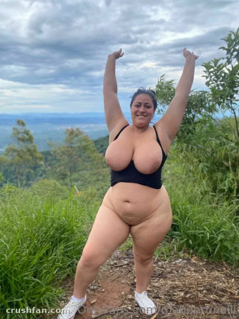 Marcela in einem schwarzen Crop-Top mit erhobenen Armen auf einem grasbewachsenen Hügel unter einem bewölkten Himmel