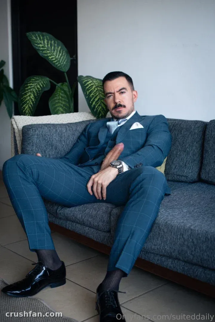Creator in a teal three-piece suit seated on a grey sofa beside an indoor plant.