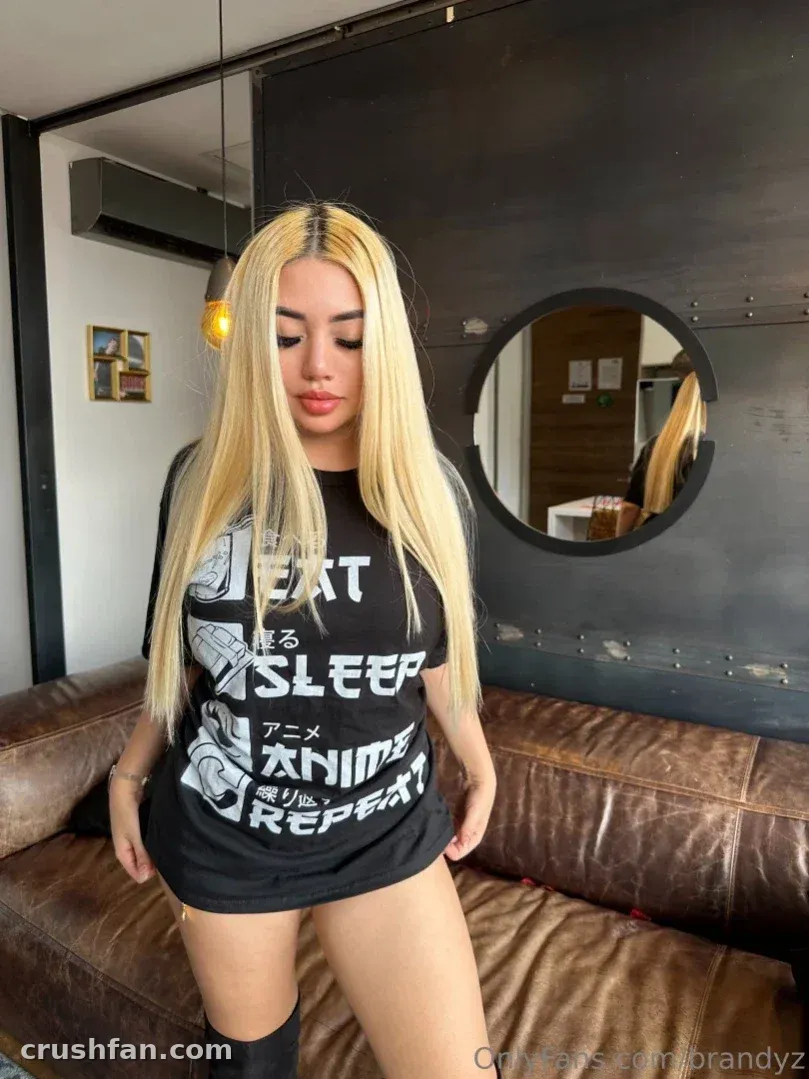 Brandy Bunny Bunz wearing a black graphic tee while standing next to a brown leather sofa indoors.