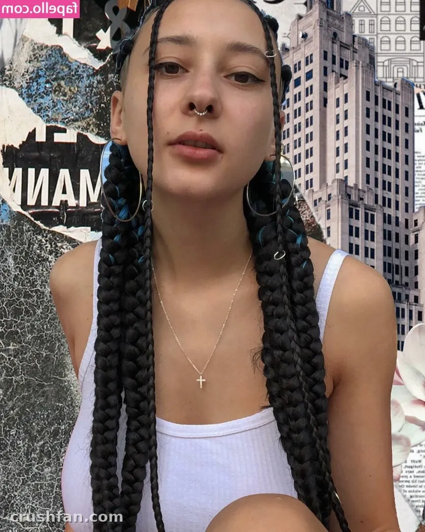 𝐋𝐢𝐥𝐚 in a white tank top with long dark braids, standing before a collage of posters and a city building.