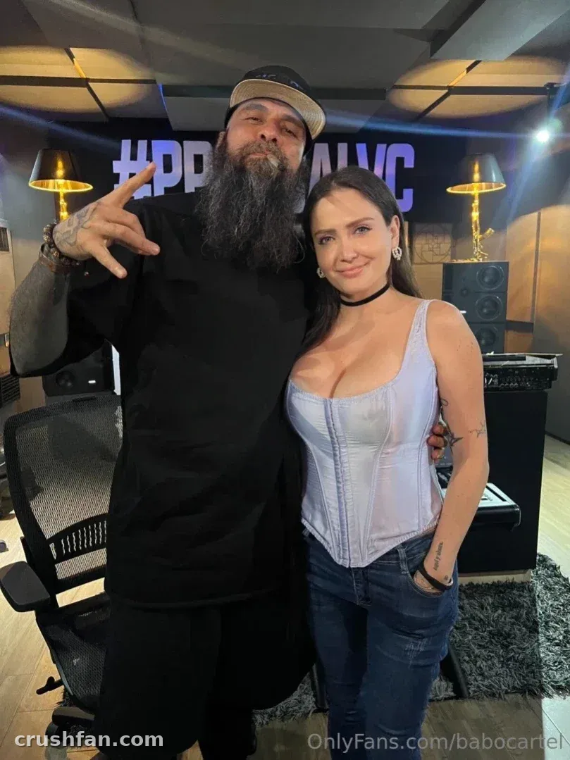 Babo Cartel de Santa mit schwarzer Kappe und dunklem Hemd neben einer Frau in einem lila Korsett-Top in einem Studio.