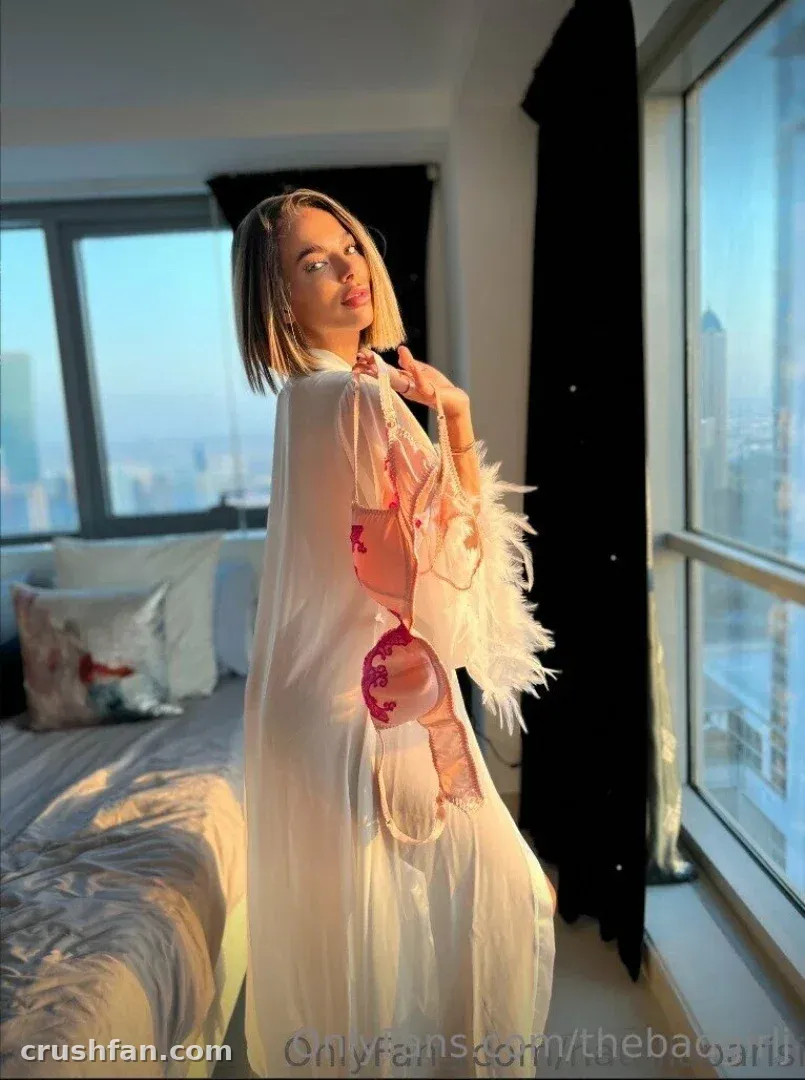 Creator in a sheer white robe with pink details standing by a window in a bedroom with a city view.