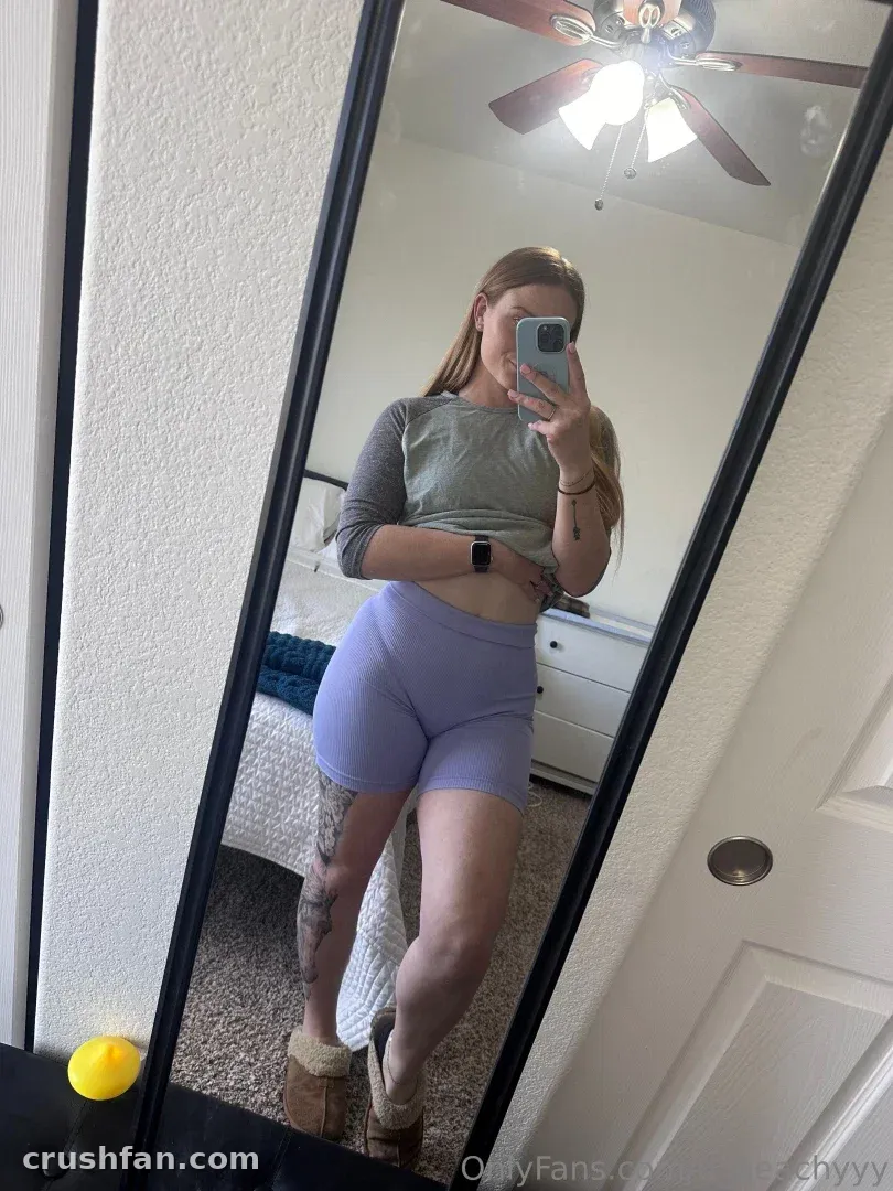 Dee Jacobs com top cropped cinza e shorts lilás tirando uma selfie no espelho em um quarto.