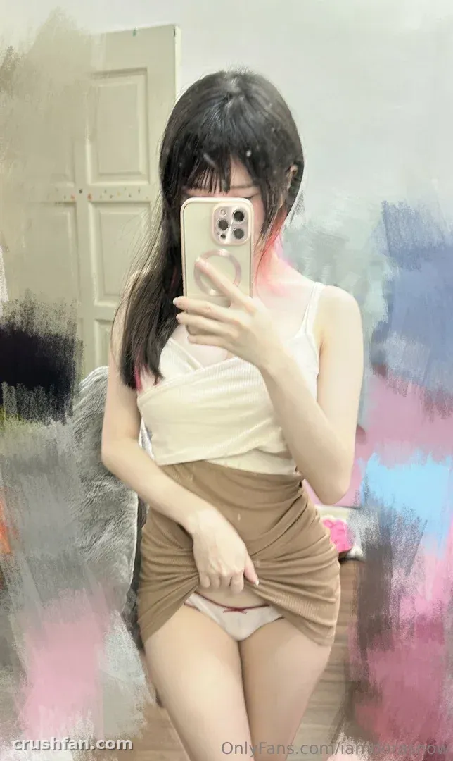 Sally Dorasnow com um top creme e saia bege tirando uma selfie no espelho em ambiente interno