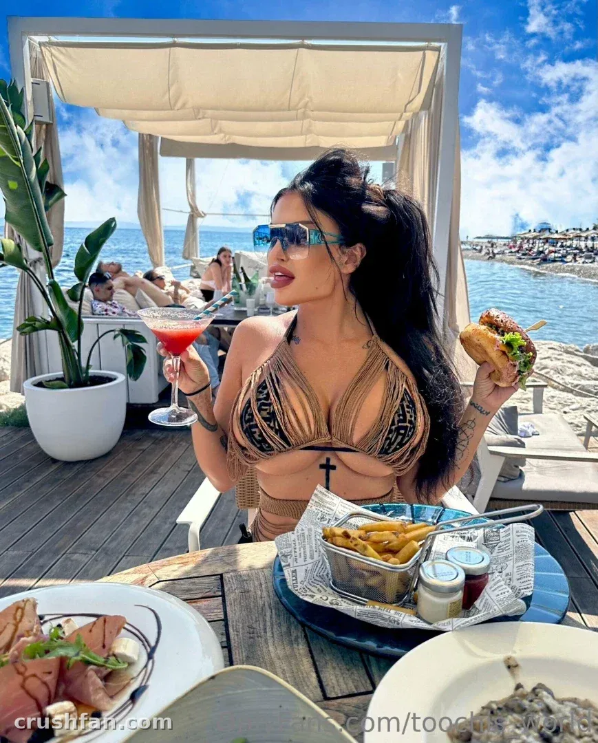 Toochi Kash in a bikini and sunglasses holding a drink and burger at a seaside cabana table.
