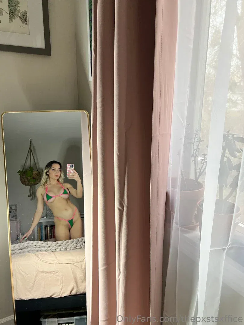 Maizie de biquíni verde tirando uma selfie no espelho em um ambiente iluminado com cortinas rosa