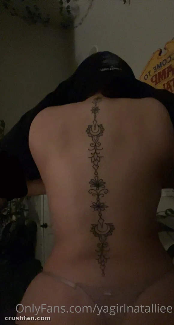 Natalie seen from behind showing a long ornamental spine tattoo in low light
