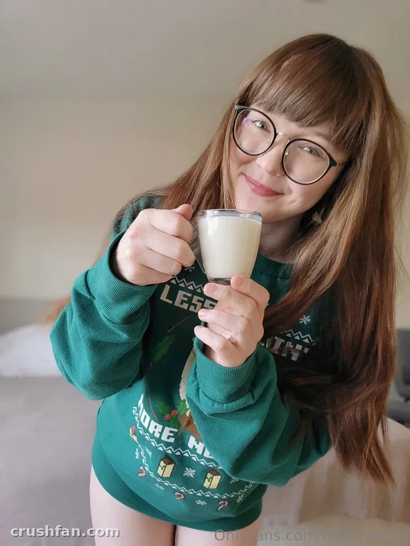 Mira ♡ in a green festive sweater and glasses holding a glass of milk indoors.