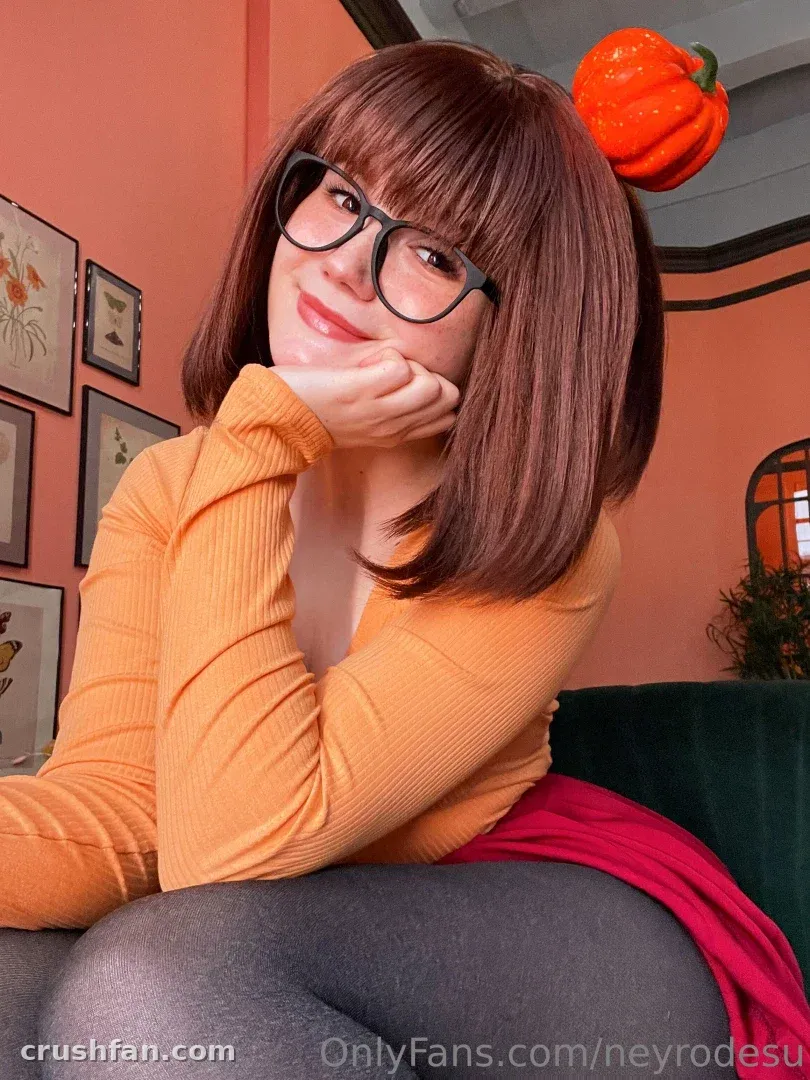 Ney in an orange sweater and glasses, chin on hand, pumpkin headband, coral wall behind