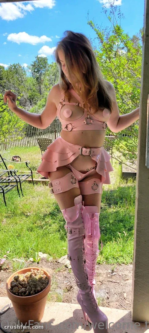 Creator dans une tenue rose à lanières et de hautes bottes posant sur un porche ensoleillé avec un jardin derrière elle