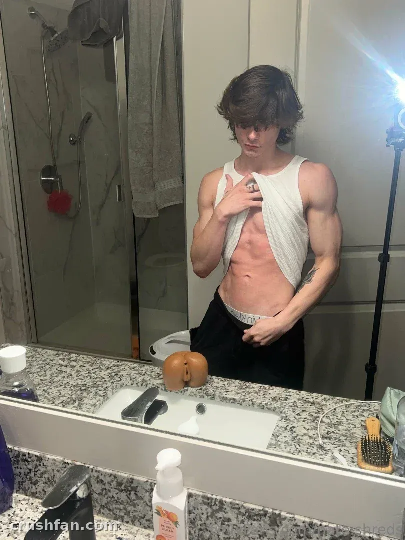 NATHAN lifting a white tank top in front of a bathroom mirror with a granite counter