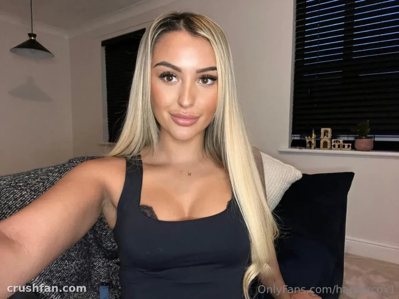 Harper in a black tank top taking a selfie on a grey sofa indoors