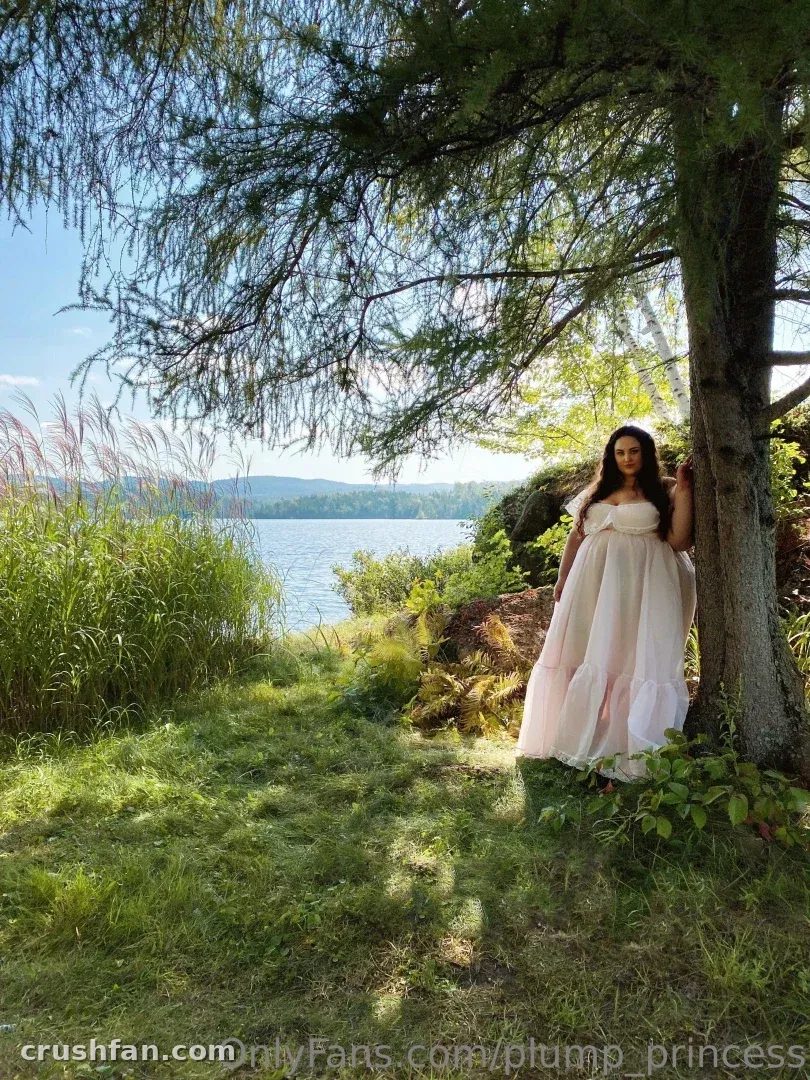 Plump Princess in a long gown standing by a tree near a lake