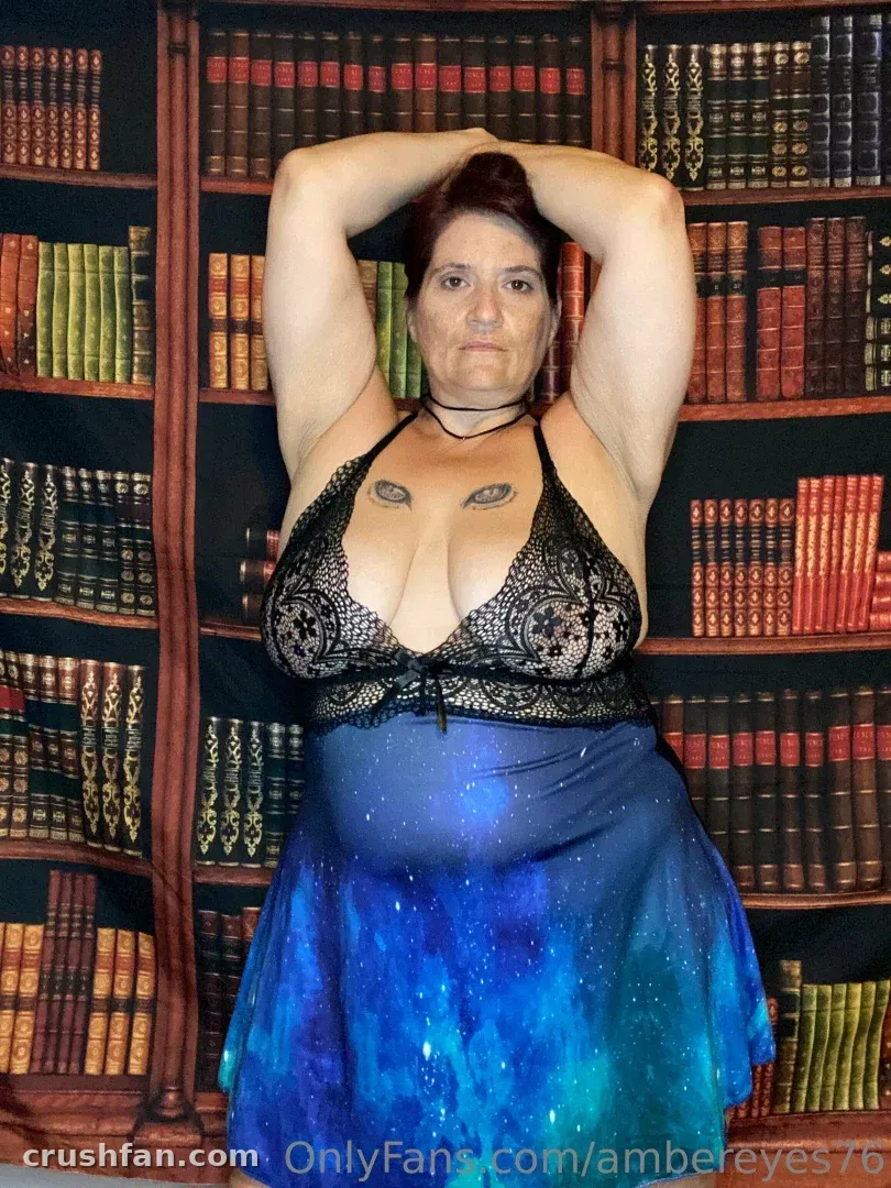 Amber Eyes dans un haut en dentelle noire et une jupe bleue debout devant une bibliothèque