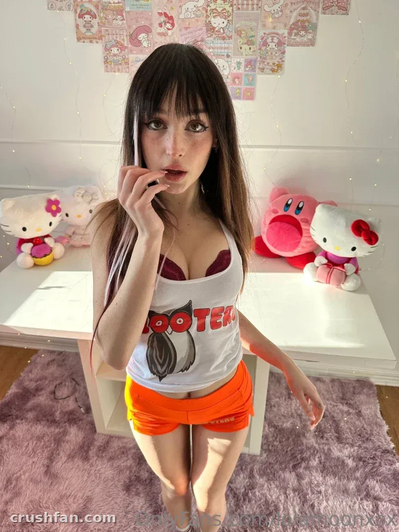 Lola in a white graphic top and orange skirt standing by a desk with plush toys
