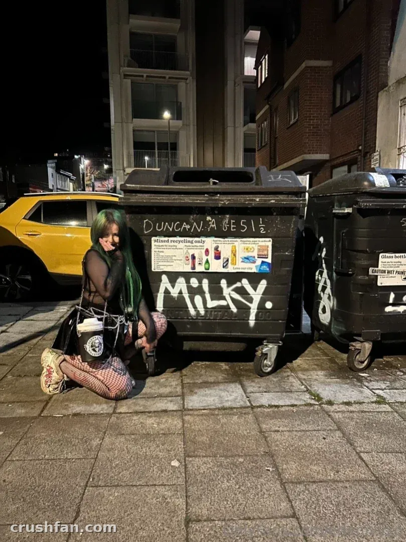 Eden crouches on a night sidewalk beside a graffiti-covered bin and a yellow car.