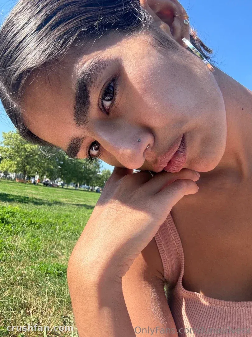 Close-up outdoor selfie of a woman in a pink top with a sunny park behind her