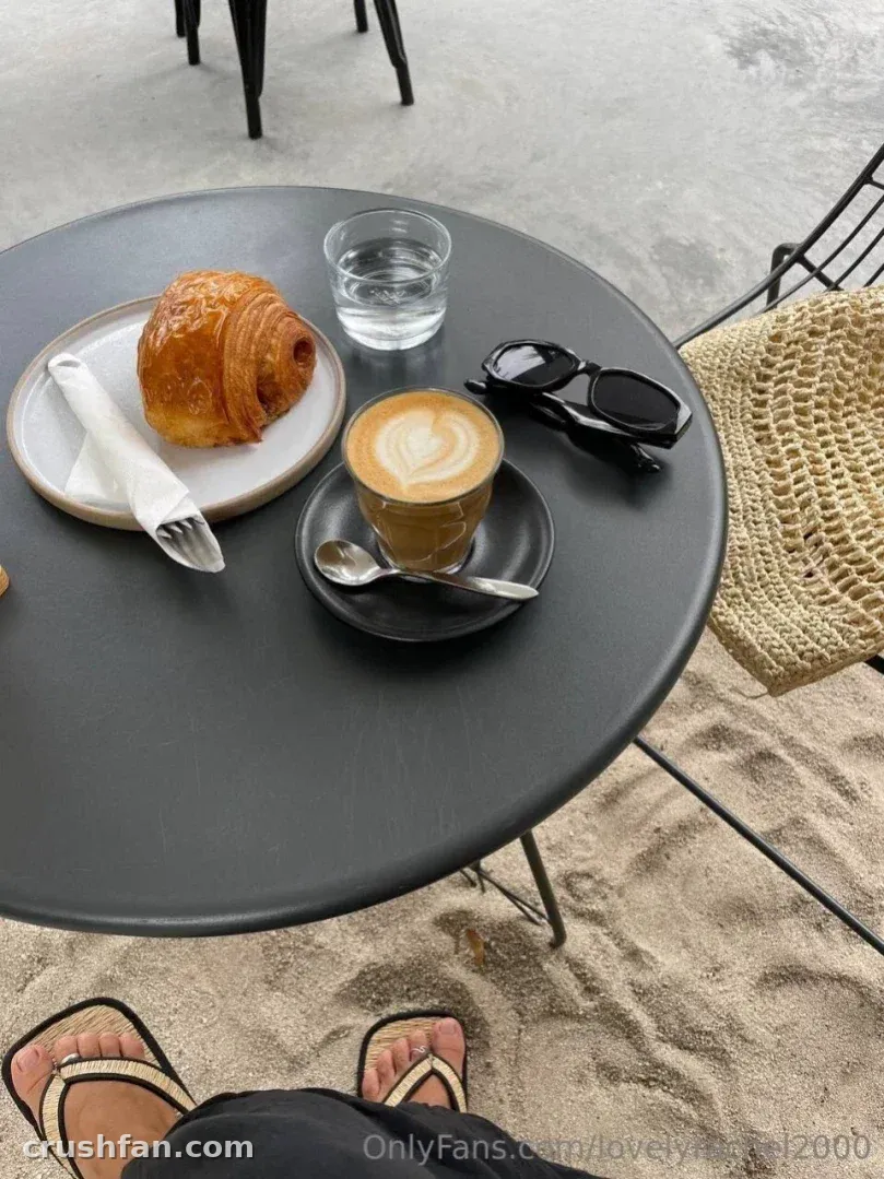 Coffee, croissant and sunglasses on a table beside sand, with feet in sandals below.